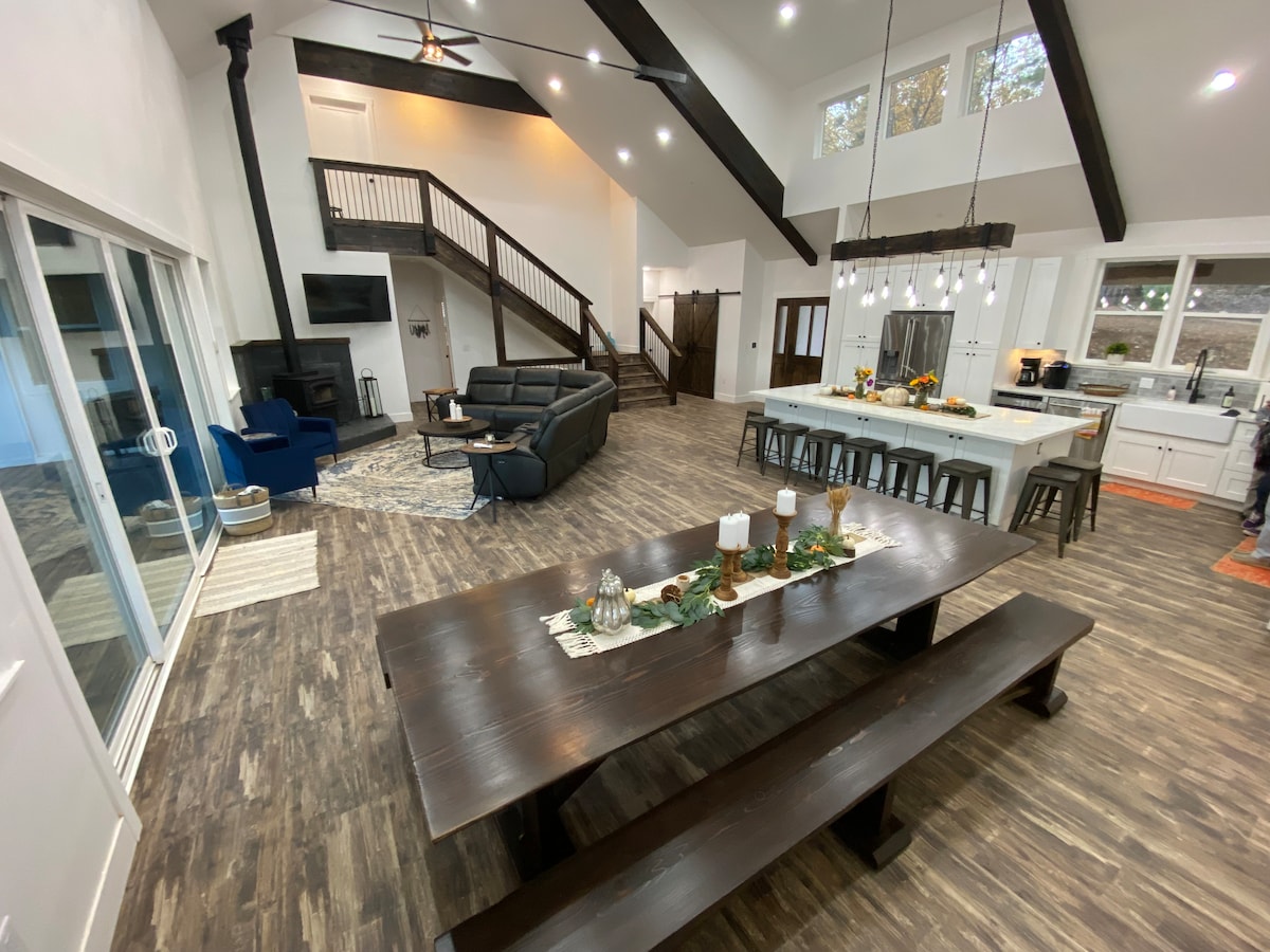 An open-concept living space is highlighted by vaulted ceilings and dark wooden beams. A large dining table occupies the center, surrounded by seating areas. Large glass doors allow natural light and a view of the outdoor area, enhancing the room's spacious feel.