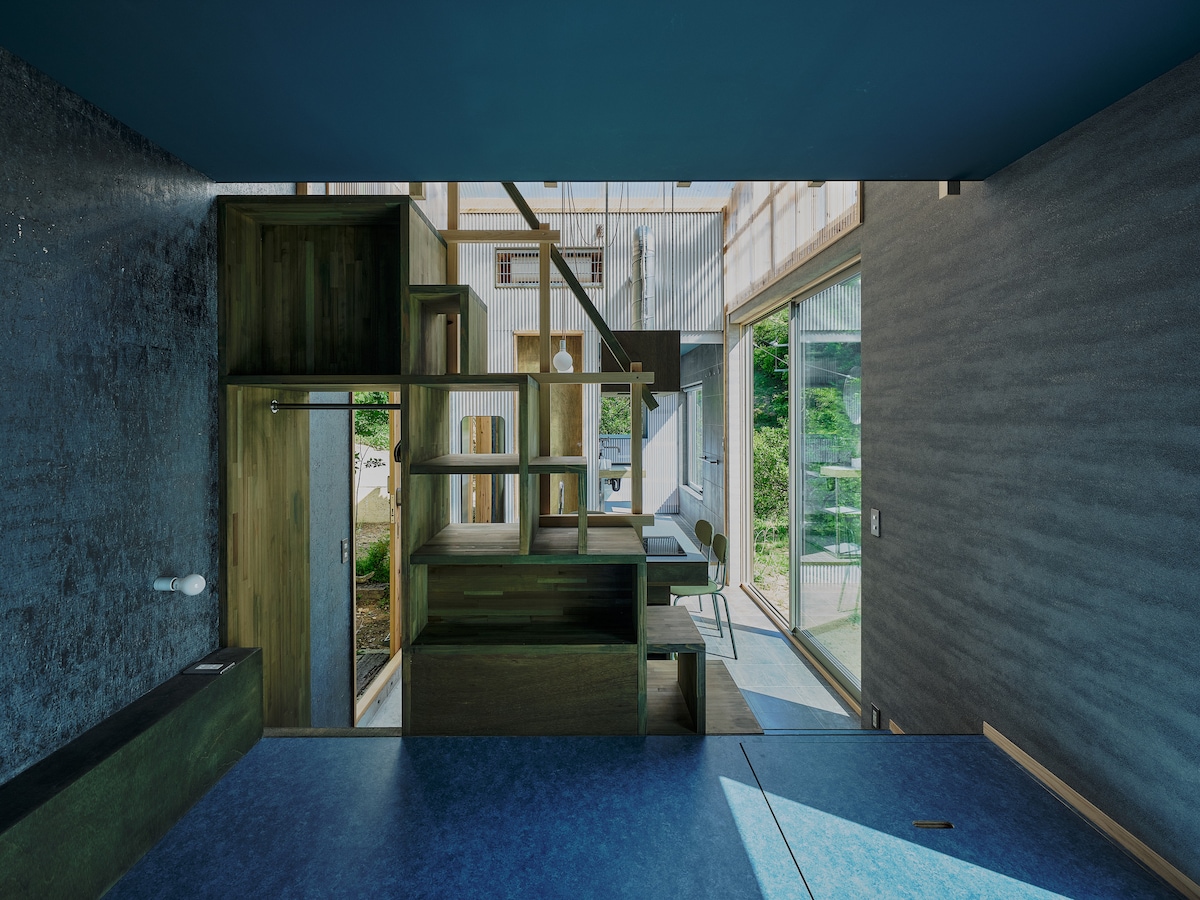 A modern interior is showcased with various wood structures creating a unique, multi-level design. Large windows allow natural light to fill the space, with a view of greenery visible through the glass. A mix of textures in the walls adds depth to the room.