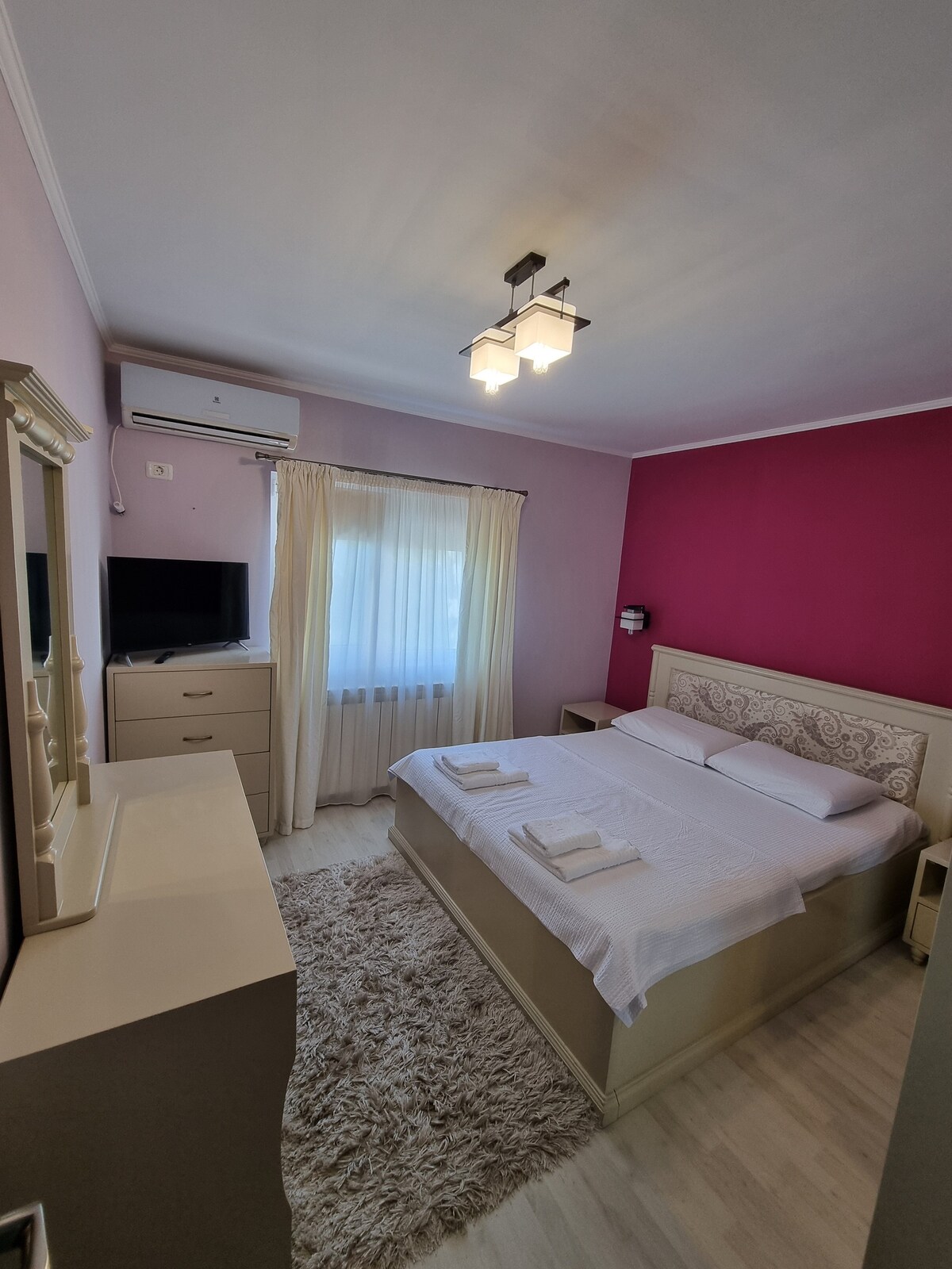 A well-appointed bedroom features a spacious bed with light bedding against a pink accent wall. Natural light enters through a white curtain-draped window. A light-colored dresser and a wall-mounted television complete the space, which is complemented by a soft area rug beneath.