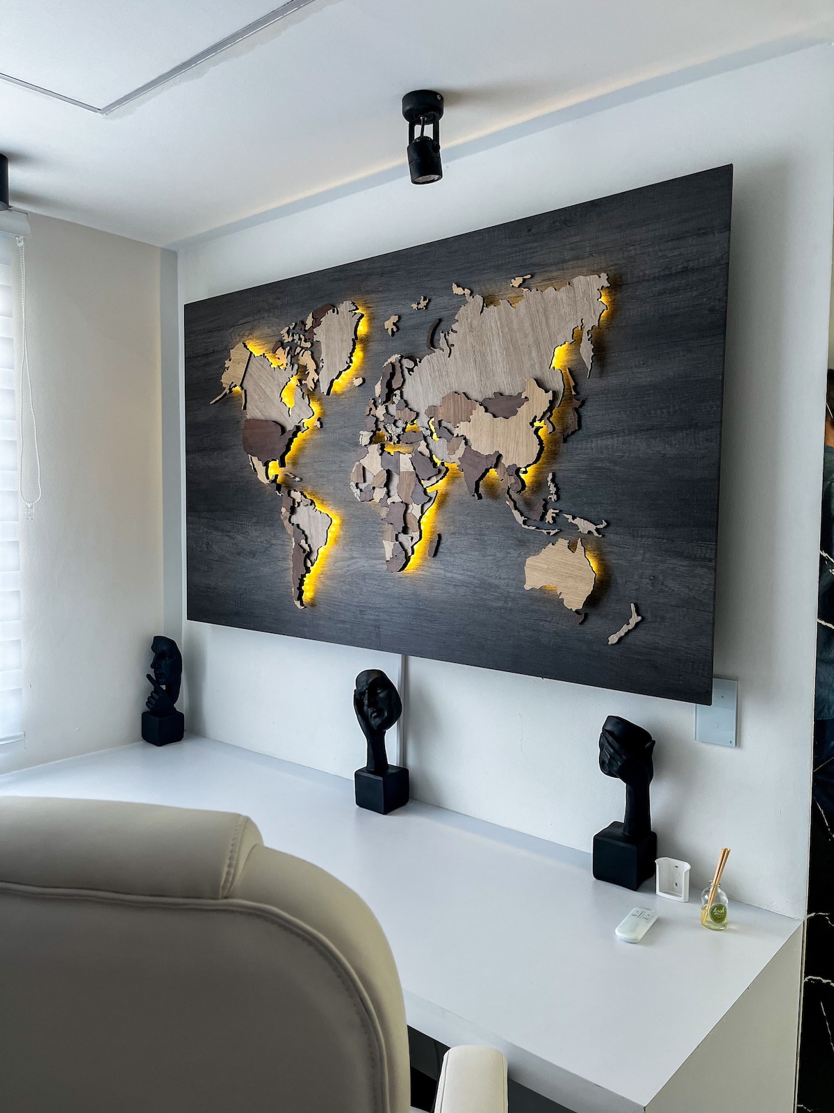 A detailed wooden map of the world is showcased on a feature wall, bordered by soft, glowing lights. Underneath, a sleek desk with two sculpted figures provides an artistic touch. Natural light filters through partially closed blinds, creating a bright and inviting workspace.
