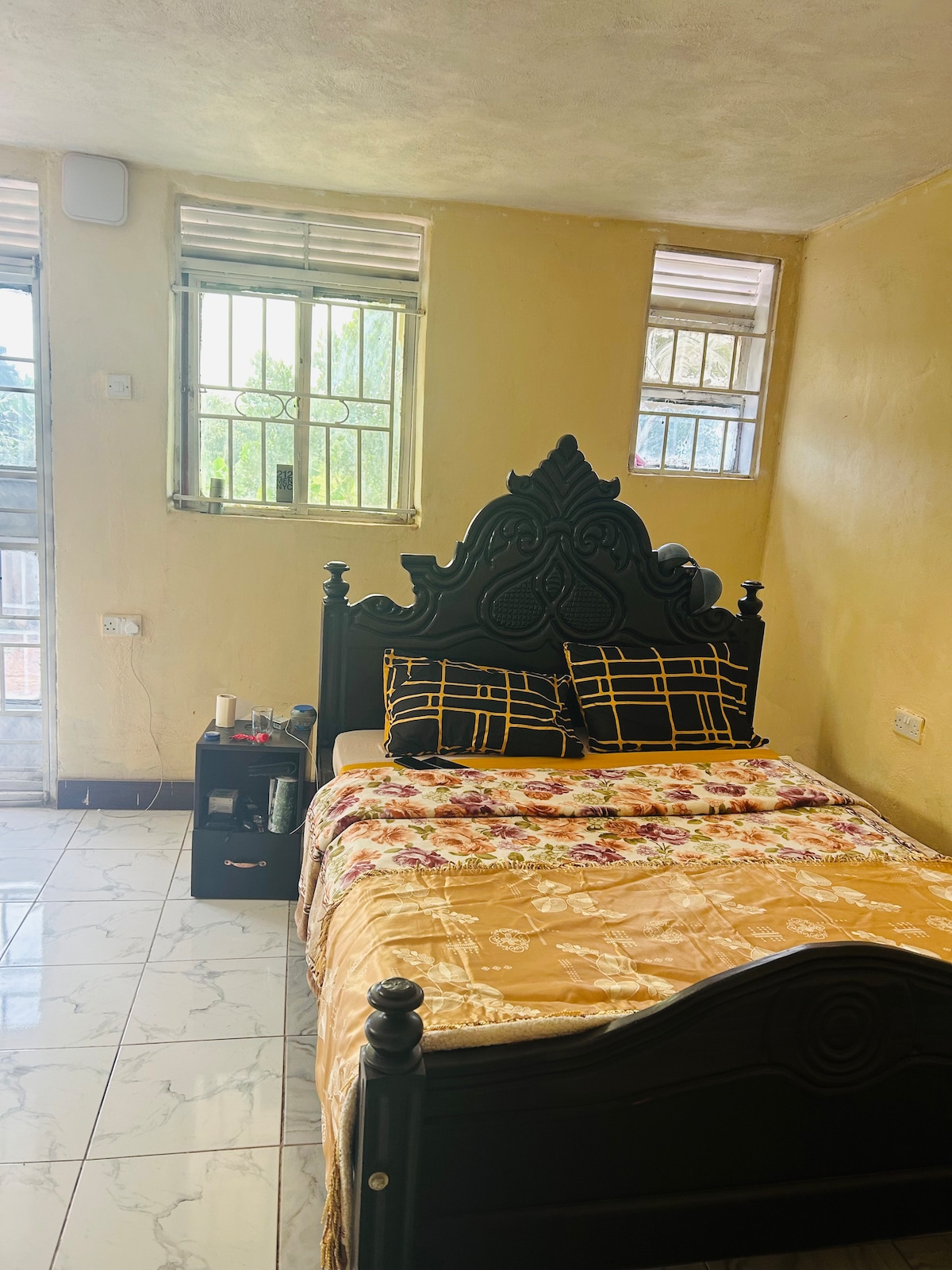 A cozy bedroom features a decorative black bed frame with a plush yellow and floral patterned bedding. A bedside table holds essential items. Natural light filters through two windows, illuminating the room's light-colored walls and tiled floor.