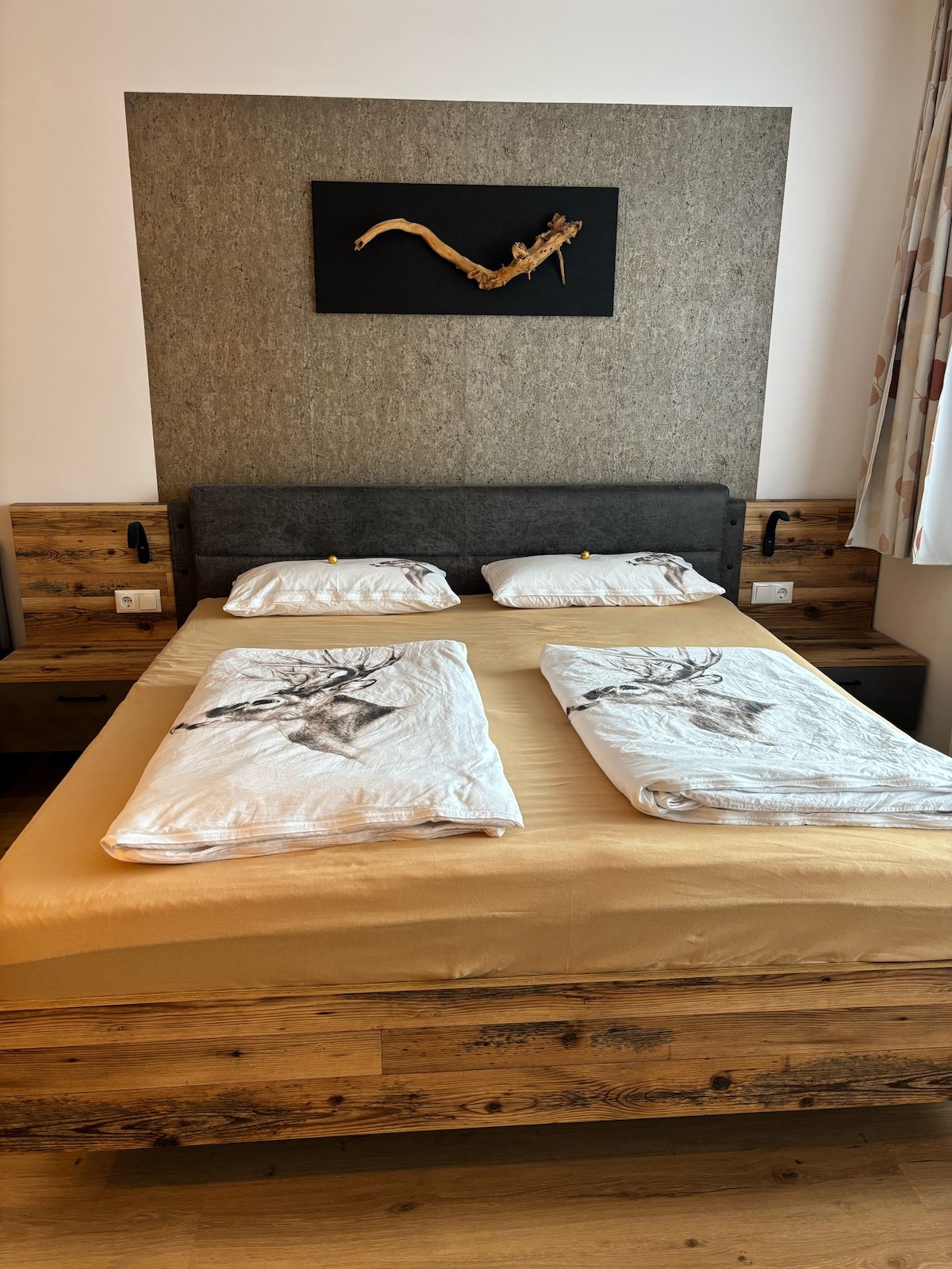A spacious bed is presented with light-colored bedding featuring nature-inspired prints. The headboard is designed with a textured gray wall, complemented by dark wood nightstands on either side. Natural wood elements are integrated into the room for a cohesive look.