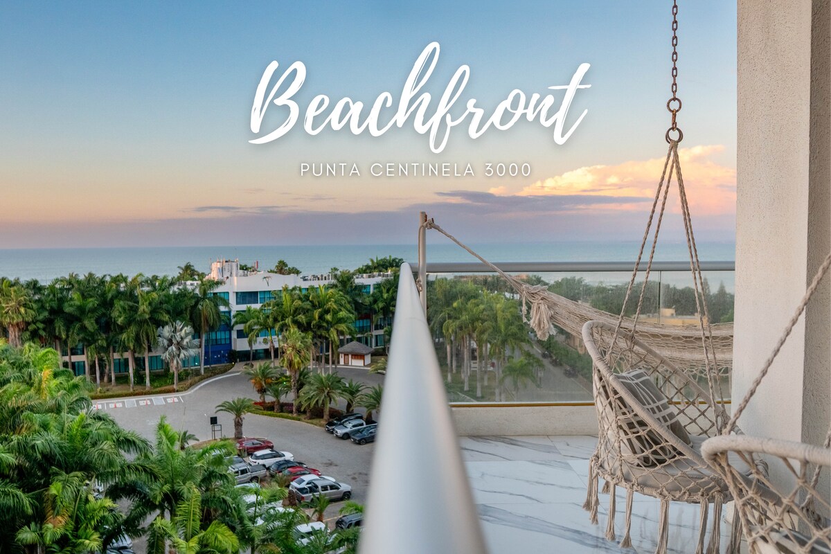 A balcony view overlooks lush greenery and a distant ocean horizon, with a hanging hammock positioned to invite relaxation. The sunset casts soft colors across the sky, enhancing the tranquil ambiance.