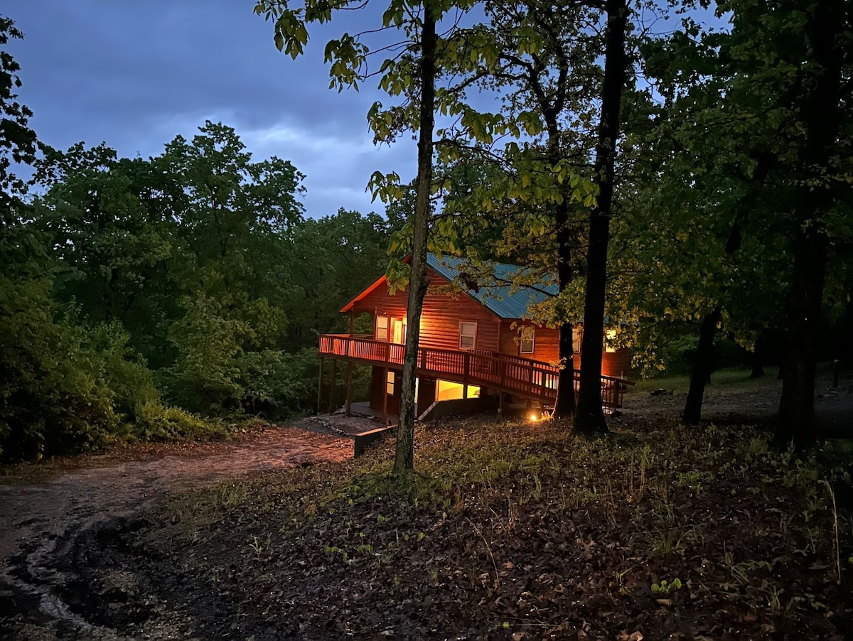 A cozy cabin nestled among tall trees is illuminated warmly in the evening. The porch extends toward a dirt pathway lined with natural foliage, creating a peaceful setting that blends into the surrounding woods.