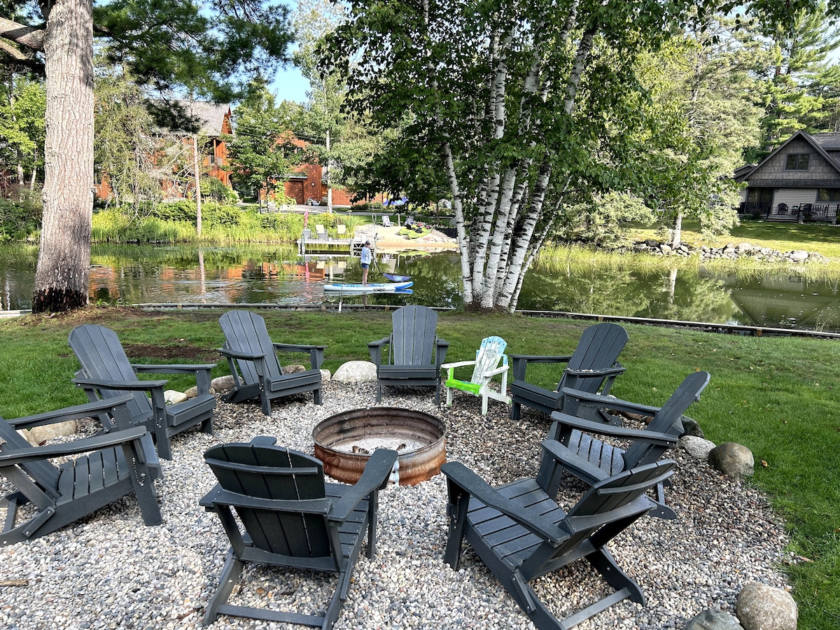 A circular arrangement of dark wooden chairs surrounds a firepit, set on a gravel area. Trees provide shade, and the water reflects the surrounding greenery. Nearby, a dock is visible with recreational equipment, enhancing the outdoor setting.