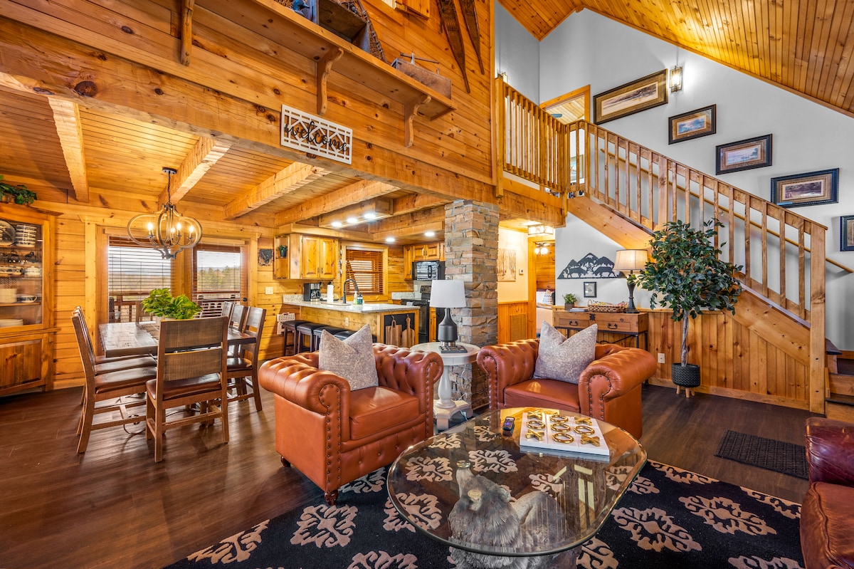 The open-concept living space features warm wooden beams and walls. A seating area with leather furniture surrounds a glass coffee table. A dining area is visible in the background, leading to the kitchen with modern appliances, while a staircase ascends to the upper level.