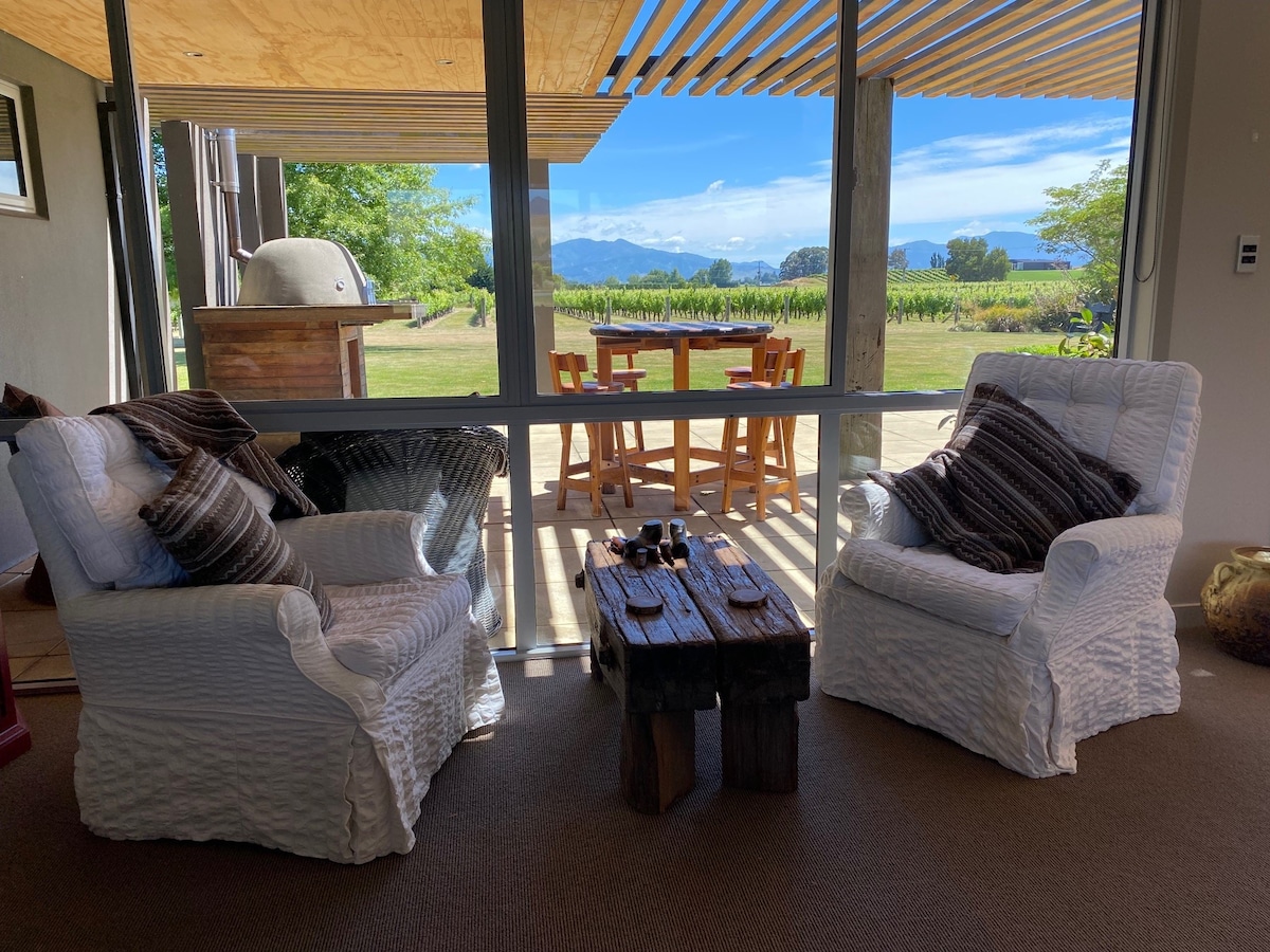 Two white, textured armchairs are positioned around a rustic wooden coffee table. A view of the outdoor dining area and vineyard is visible through large glass doors, with distant mountains creating a serene backdrop. Soft pillows add comfort to the seating.