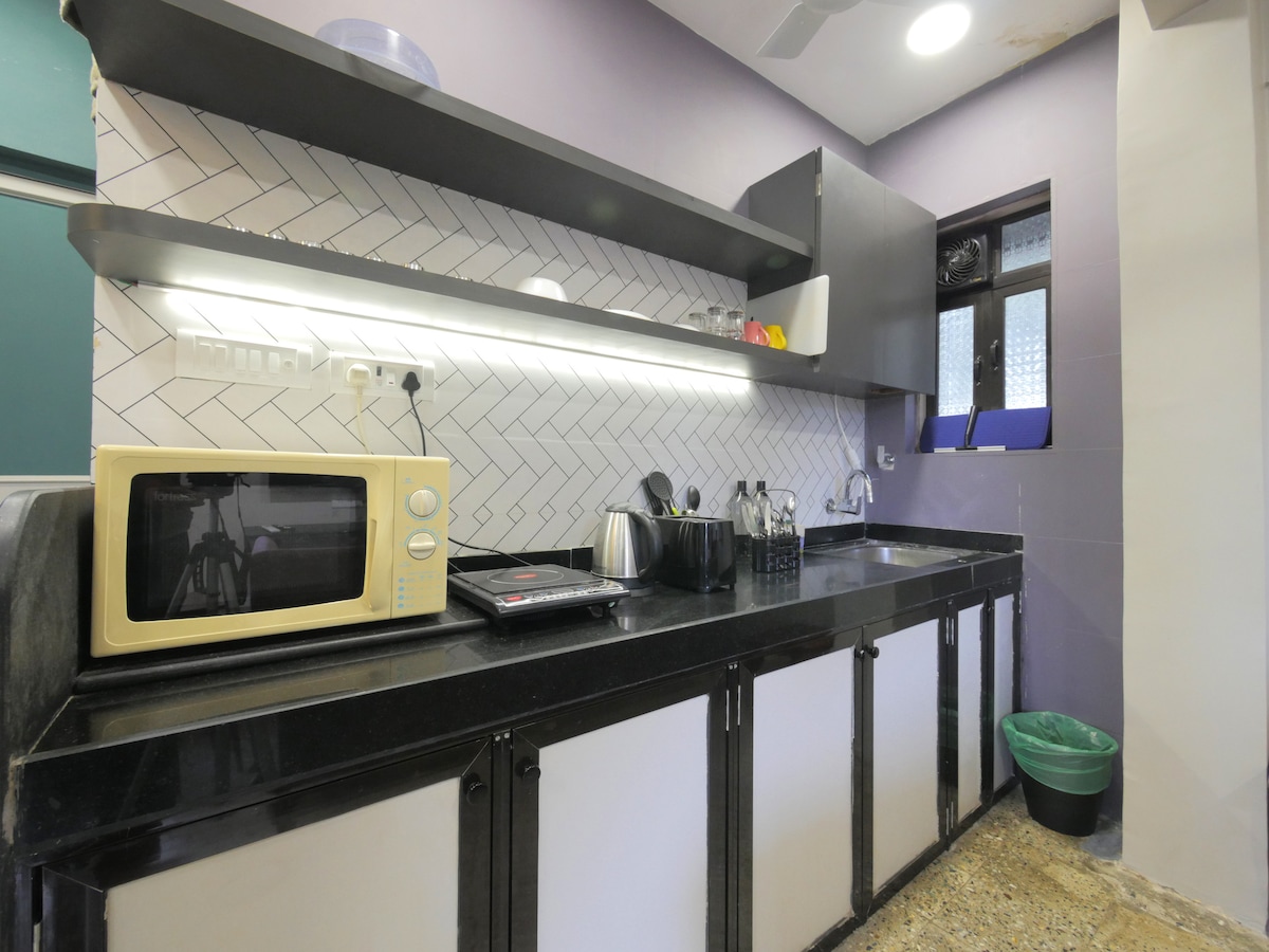 A modern kitchen is presented with sleek black countertops and a herringbone-patterned backsplash. Essential appliances include a microwave and a stovetop, with utensils and cookware neatly arranged on the shelves above. Natural light filters through a window, illuminating the workspace.