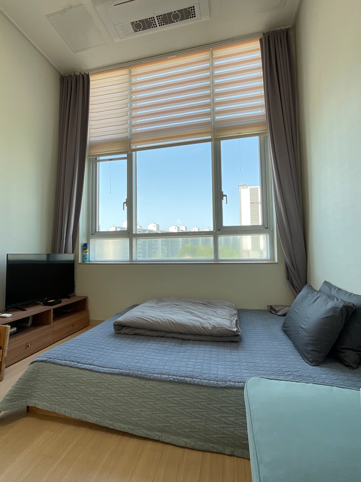 A light-filled room features a spacious bed with grey bedding and soft pillows. Natural light filters through large windows equipped with blinds. A wooden TV stand holds a television, and a small seating area is visible in the corner.