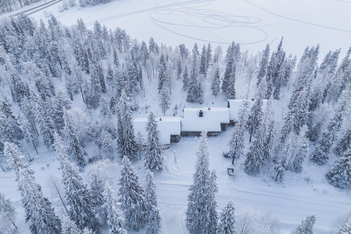 An aerial view captures a serene landscape blanketed in snow. A wooden cabin, situated amidst tall evergreen trees, is flanked by a frozen lake reflecting the surrounding winter scenery. The peaceful environment is complemented by the untouched snow along the pathways.