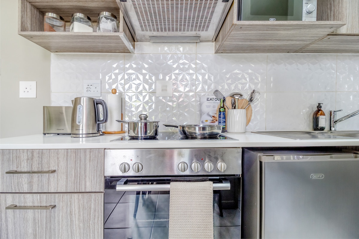 A modern kitchen showcases sleek cabinetry and a white countertop. Stainless steel appliances, including a stove, kettle, and refrigerator, are visible. A pot on the stove suggests meal preparation, while utensils and cooking essentials are neatly arranged for convenience.