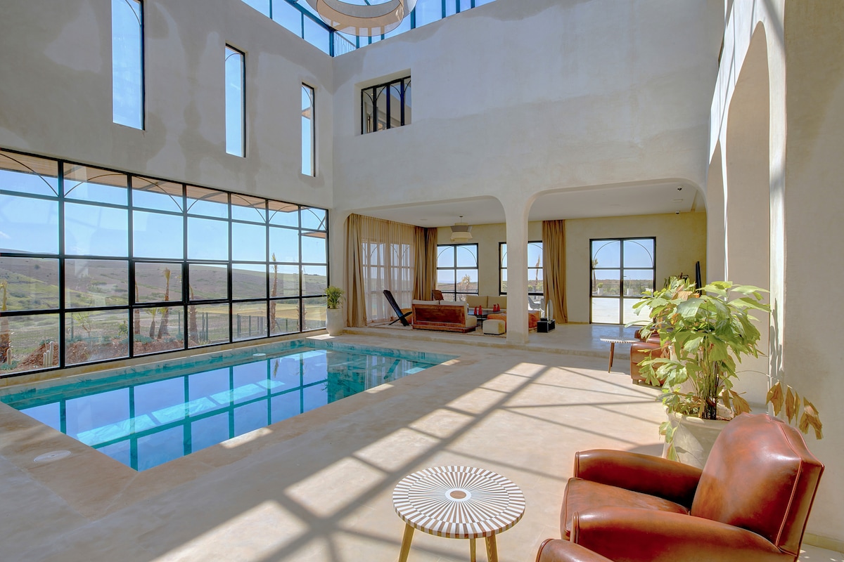 A spacious indoor area is highlighted by large windows, allowing ample natural light and offering views of the surrounding landscape. A swimming pool sits at the center, complemented by seating arrangements featuring stylish brown chairs and a decorative table.