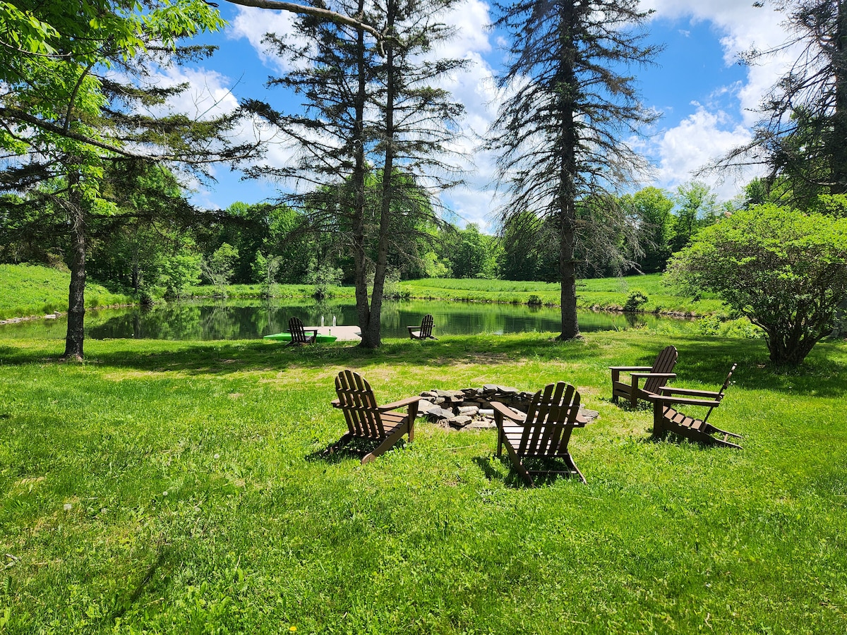 A grassy area near a pond features several wooden Adirondack chairs arranged around a fire pit. Tall trees provide shade, and the serene water reflects the clear blue sky. The landscape showcases a lush natural setting, inviting relaxation and outdoor activities.