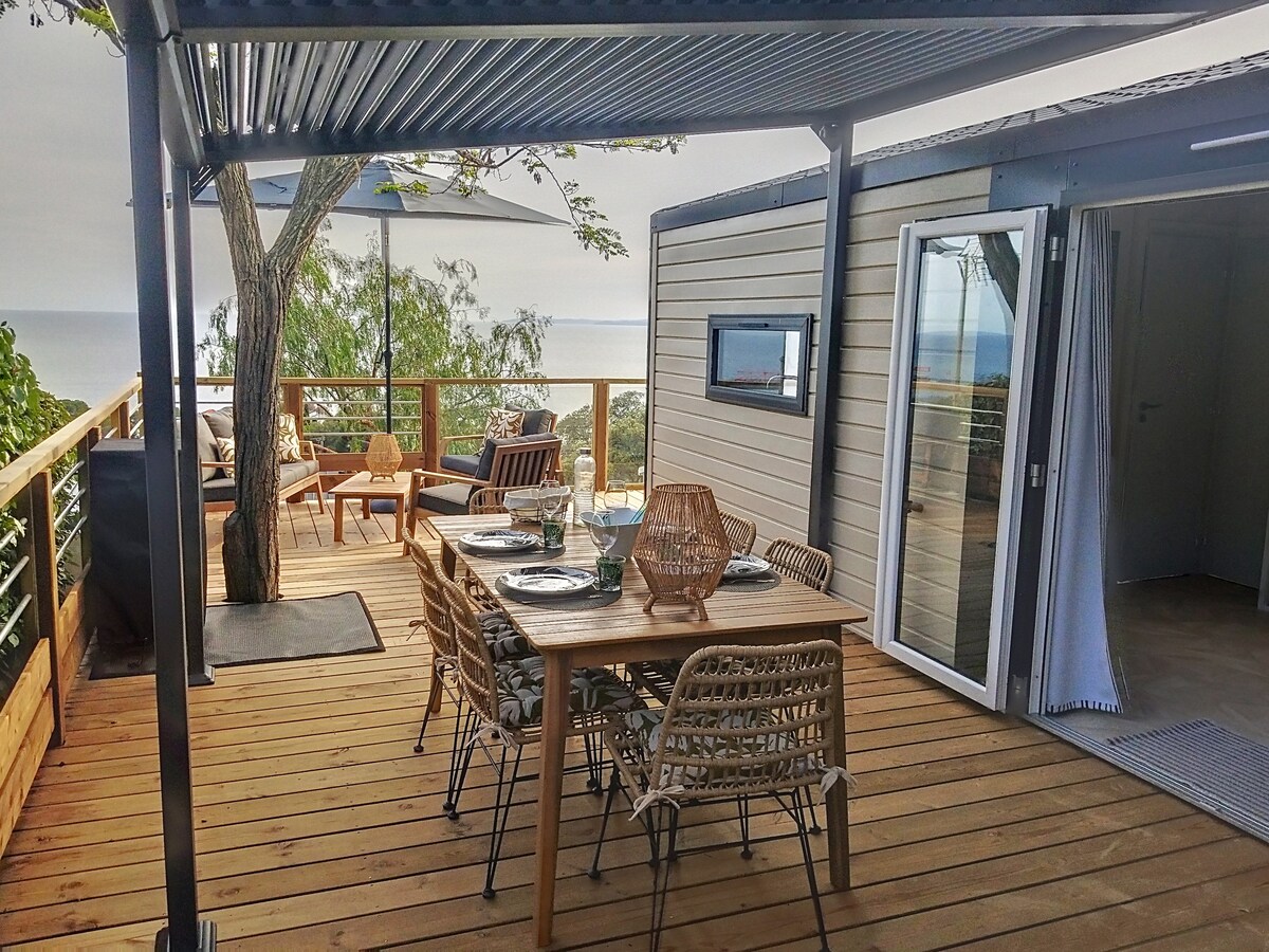A spacious terrace features a wooden dining table set with glassware, surrounded by woven chairs. A large umbrella provides shade, while views of the sea are visible in the background. A tree is situated nearby, adding a natural touch to the setting.