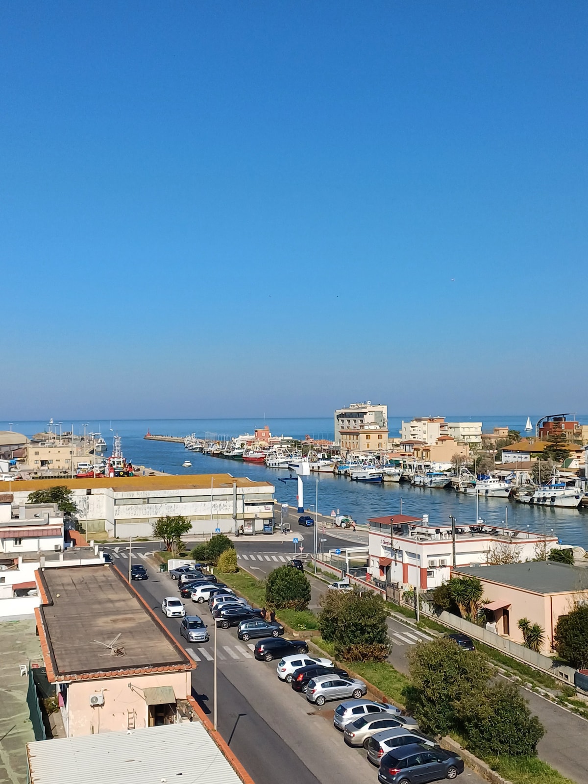 A panoramic view showcases the coastal scenery, featuring the blue sea meeting the sky in the distance. The vibrant waterfront is lined with buildings and boats, while a street filled with parked cars and greenery is visible in the foreground.