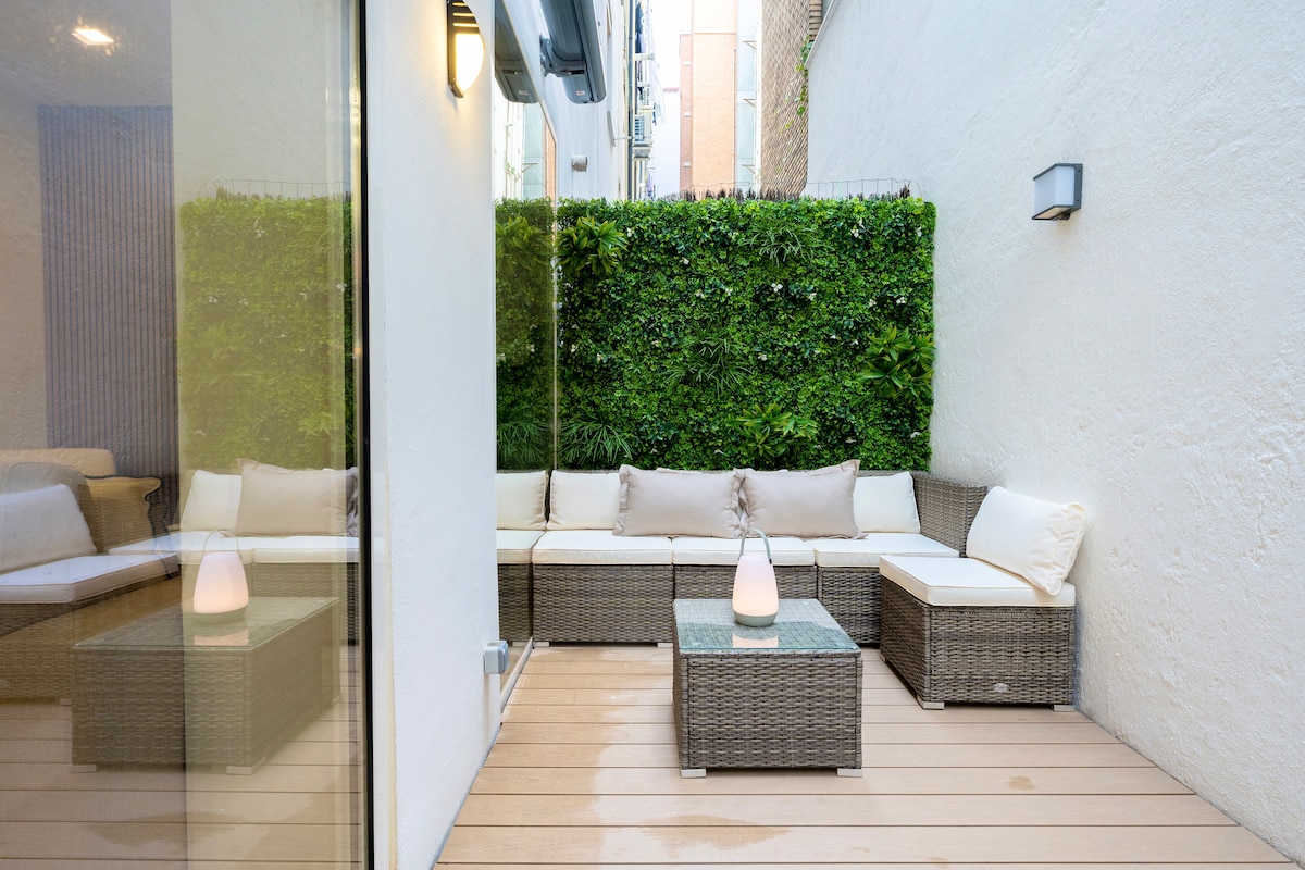 A private terrace features a cozy seating area with a sectional sofa adorned in light cushions. A glass-top coffee table rests on a wooden deck, complemented by a modern light fixture. Lush greenery provides a natural backdrop, enhancing the relaxing environment.