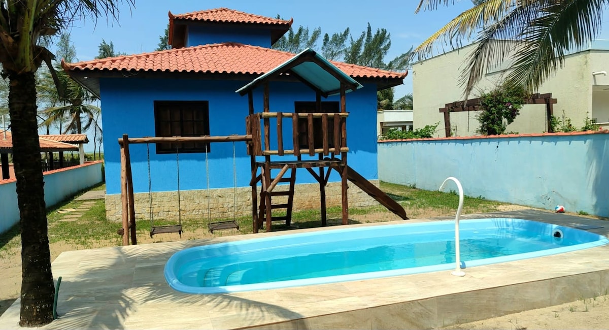 A vibrant blue house stands in a spacious yard, featuring a small swimming pool and a wooden play structure with swings. Lush greenery surrounds the area, and palm trees add to the tropical atmosphere.