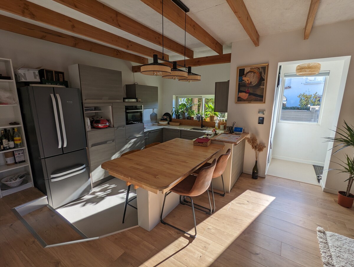 A modern kitchen features wooden beams overhead and a large dining table surrounded by four chairs. Stainless steel appliances are aligned against the wall, and a window allows natural light to brighten the space. Greenery is visible through the window, adding a touch of nature.