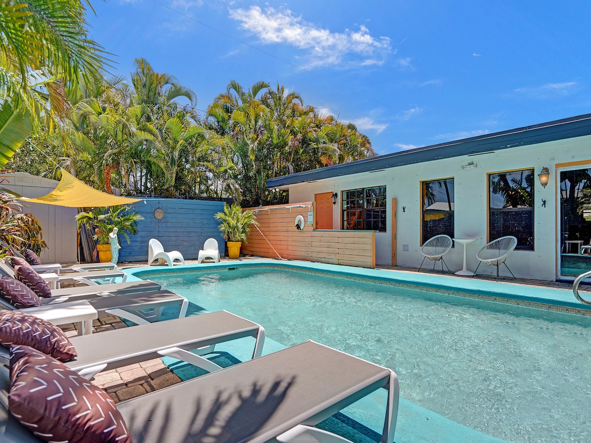 A large heated pool is surrounded by lounge chairs, inviting relaxation under the sun. Lush tropical plants border the area, creating a serene atmosphere. The pool area is complemented by an adjacent building featuring large windows, allowing for natural light to brighten the space.