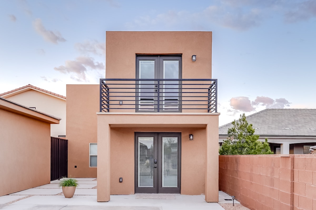 The modern exterior of the guest home features a smooth, earth-toned façade with large glass doors below a private balcony. The balcony is framed by minimalist railings, enhancing the contemporary design. A small potted plant sits on the ground, adding a touch of greenery.