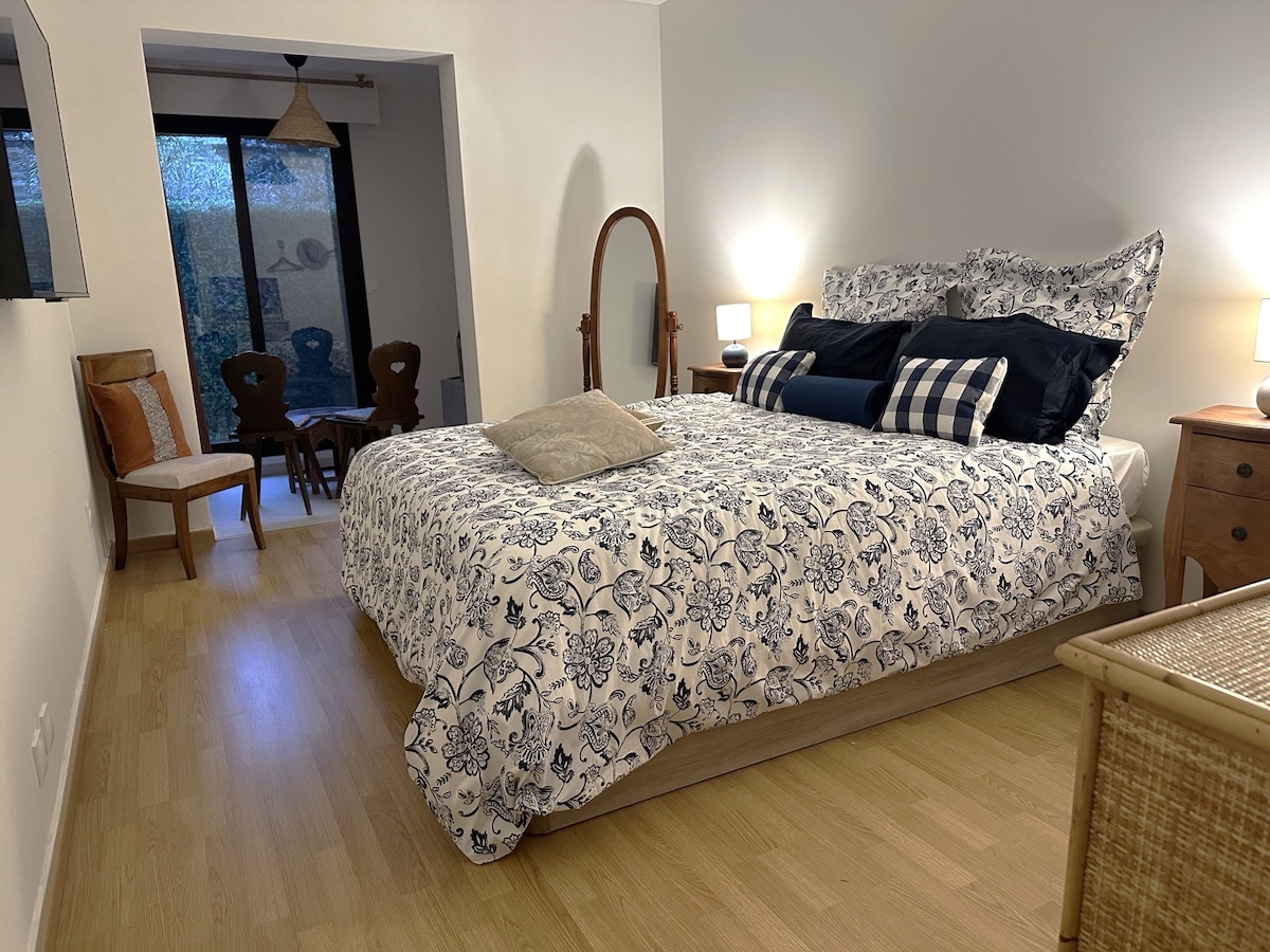A spacious bedroom is presented with a patterned bedspread and multiple decorative pillows. Soft lighting from bedside lamps creates a comforting ambiance. A mirror and seating area are visible in the background, with a large window allowing natural light to filter in.