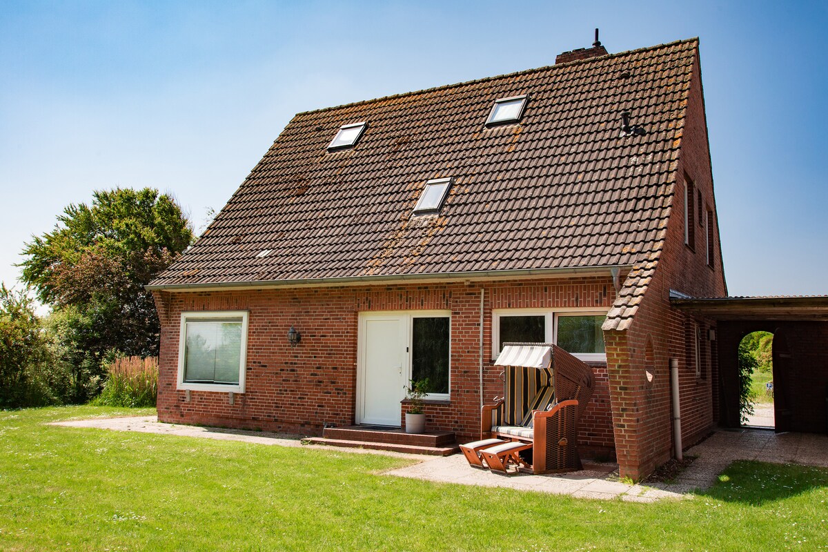 A spacious red-brick house is visible, featuring a sloped roof and multiple skylights. Large windows enhance the facade, with one window framed by a potted plant. A comfortable outdoor seating area is positioned in front, surrounded by a well-maintained green lawn.