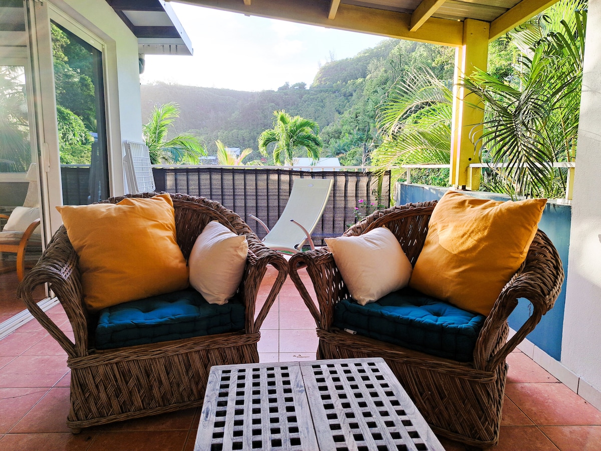 Two woven rattan chairs are positioned on a tiled terrace, each adorned with orange and blue cushions. A small coffee table is placed between them, while greenery and hills are visible in the background, enhancing the relaxing environment.