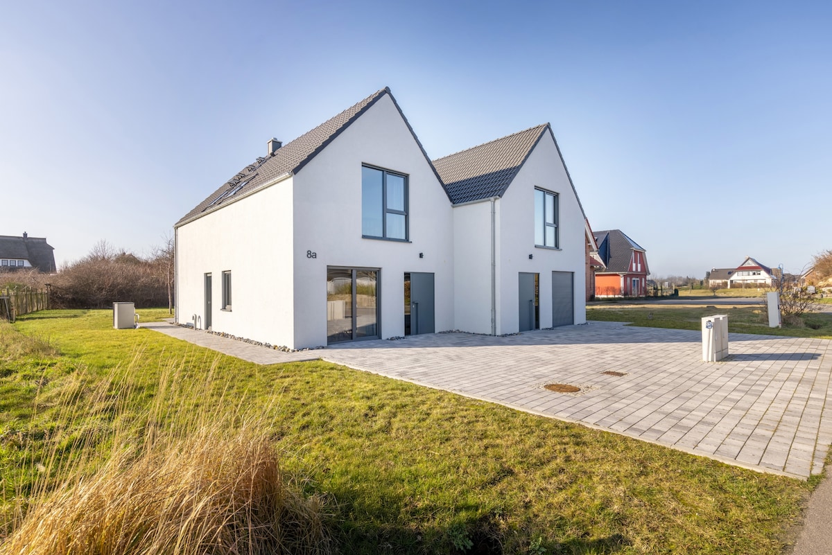 A spacious two-story house is depicted, featuring a white exterior and large glass doors. The wide paved patio area provides ample outdoor space, complemented by surrounding greenery. The landscape includes minimalistic landscaping with grass and small shrubs, enhancing the tranquil setting.
