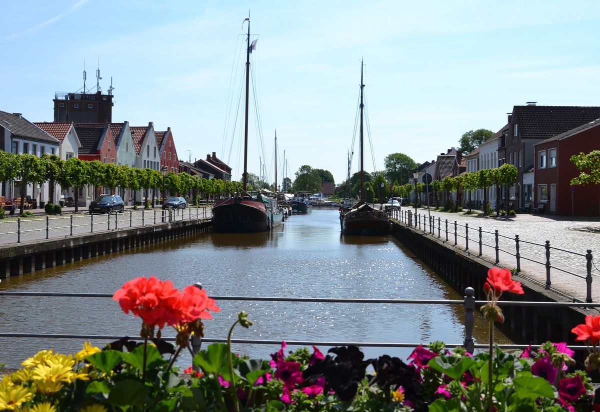 A serene view of a harbor lined with charming buildings. Two boats are moored in the calm water, while colorful flowers border the foreground. Trees are positioned along the walkway, contributing to the peaceful atmosphere of the area.