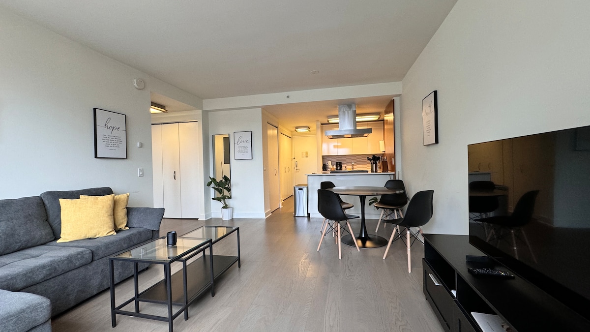 A modern living area features a comfortable gray sofa adorned with yellow accent pillows, paired with a sleek coffee table. The bright open space transitions to a streamlined kitchen view, highlighted by stainless steel appliances and ample cabinetry, all illuminated by natural light.