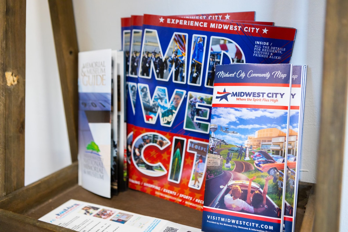 A wooden display holds various brochures and maps promoting Midwest City. Brightly colored covers showcase local attractions and events, inviting guests to explore the area. The arrangement provides easy access to information for planning activities during their stay.