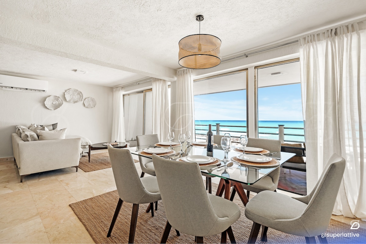 A dining area is featured, showcasing a glass table surrounded by six neutral-toned chairs. Large sliding glass doors provide a view of the coastline, while light curtains delicately frame the windows. A cozy seating area is located nearby, enhancing the spacious feel.