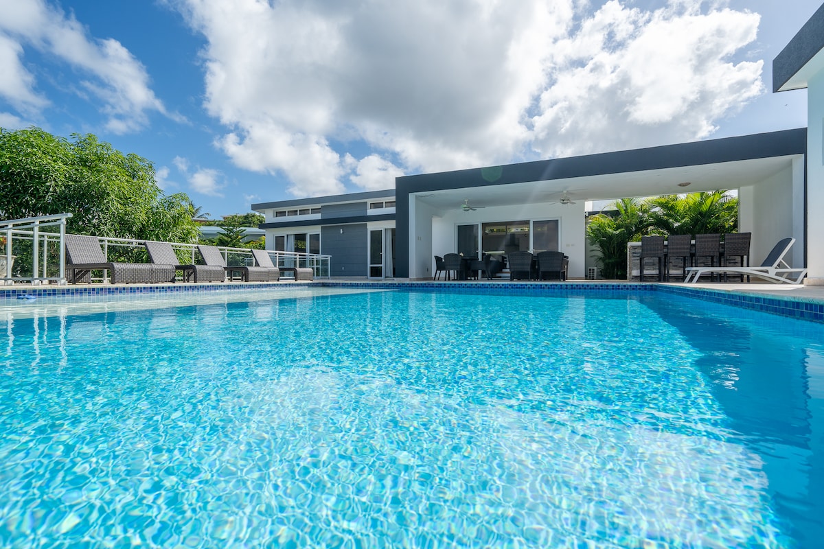 A refreshing infinity pool is framed by loungers under a clear sky, reflecting the sunlight. The modern villa is seen in the background, featuring an open terrace with dining furniture and lush greenery surrounding the area.