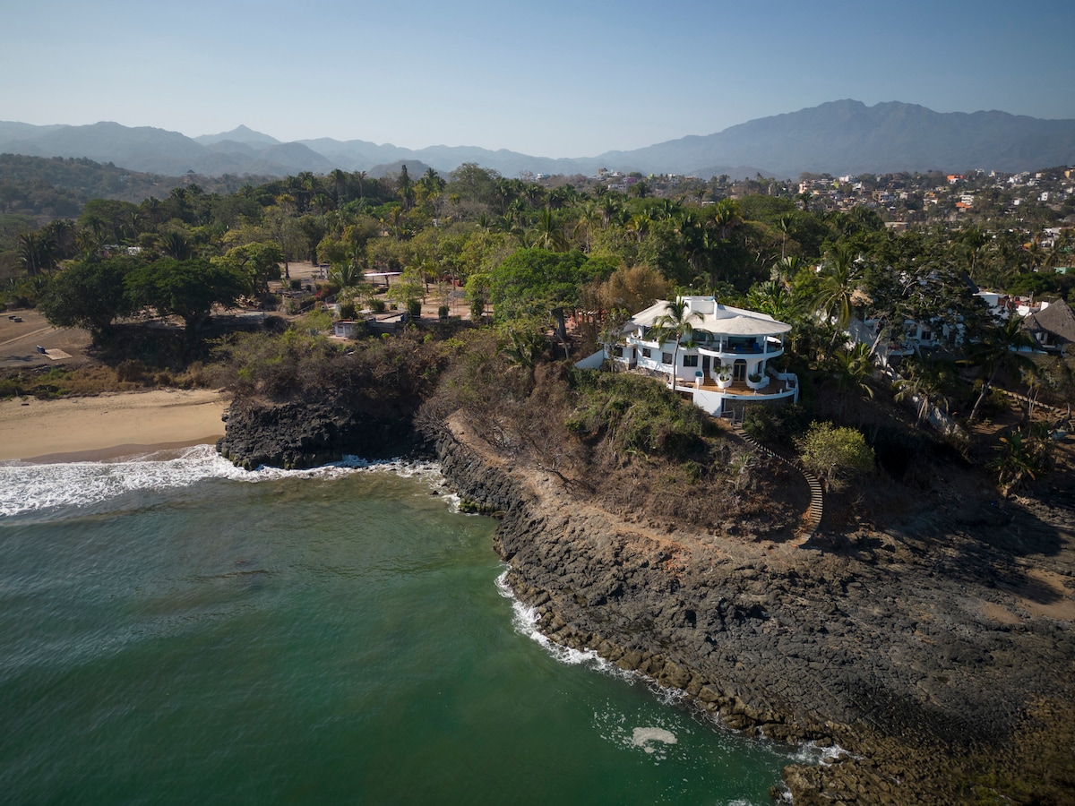 Aerial view showcases the Casa Mirador positioned on rocky volcanic cliffs, overlooking the shimmering waters of Jaltemba Bay. Lush greenery surrounds the home, with sandy beach and distant mountains visible under clear skies.
