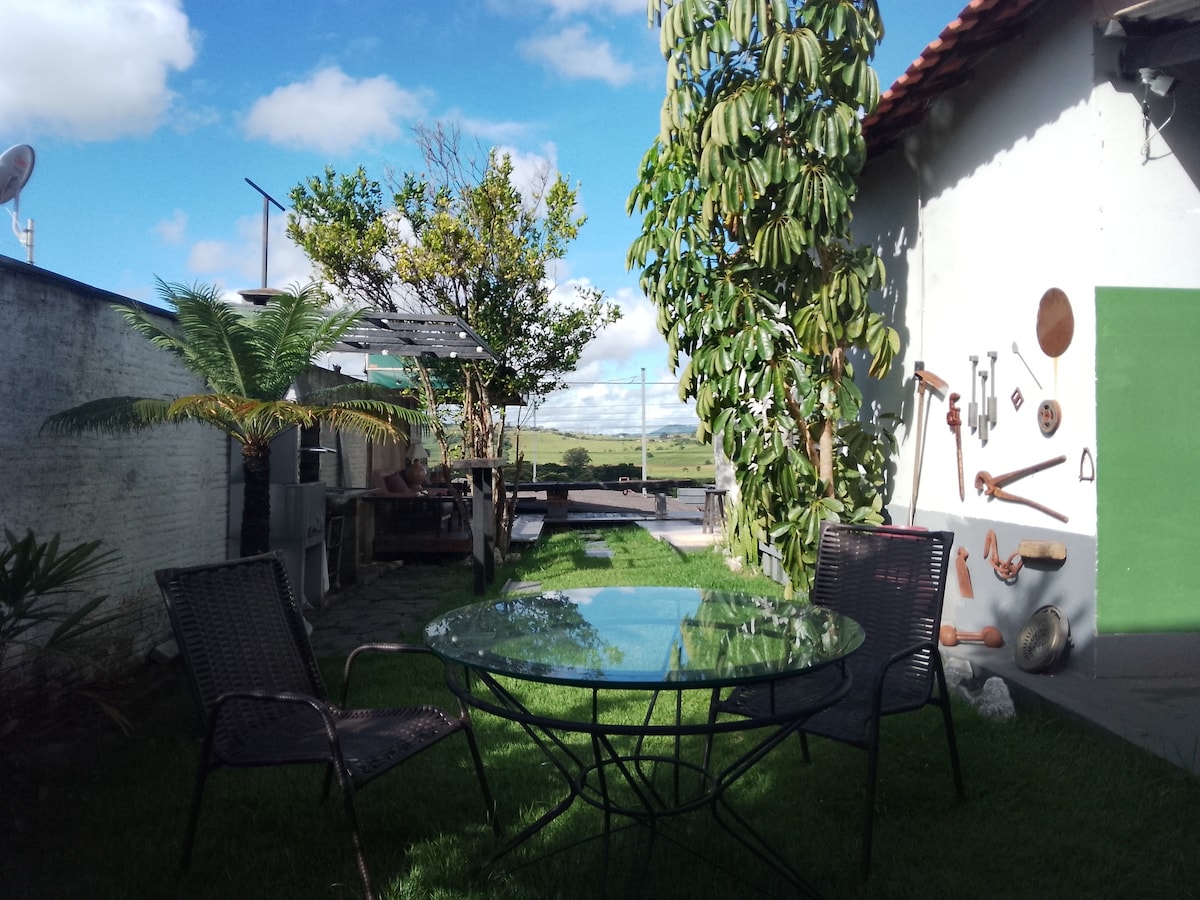 A tranquil outdoor area features a glass table surrounded by two wicker chairs on lush green grass. Nearby, various plants add natural beauty, while a wall displays an assortment of artistic wooden tools. A scenic view of the countryside is visible beyond the space.