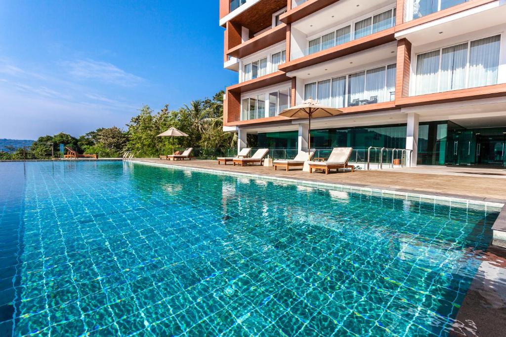 A serene outdoor swimming pool reflects the clear blue sky and is surrounded by sun loungers and umbrellas. The edges of the pool blend seamlessly into the landscaped grounds, and the modern building with large glass windows is visible in the background.
