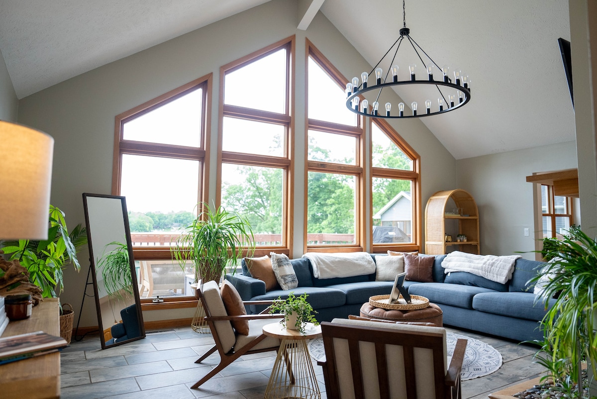 A spacious living area features large, triangular windows that frame views of the surrounding greenery. A comfortable sectional sofa is arranged around a circular coffee table, while decorative plants add a natural touch. A pendant chandelier hangs gracefully from the vaulted ceiling.