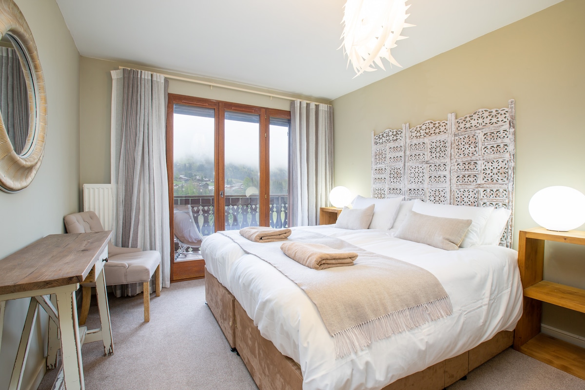 A spacious bedroom is presented with a large bed featuring light linens and decorative throws. Soft lighting is provided by round bedside lamps, and a wood accent wall is seen behind the bed. Floor-to-ceiling windows offer access to a balcony, enhancing the natural light.