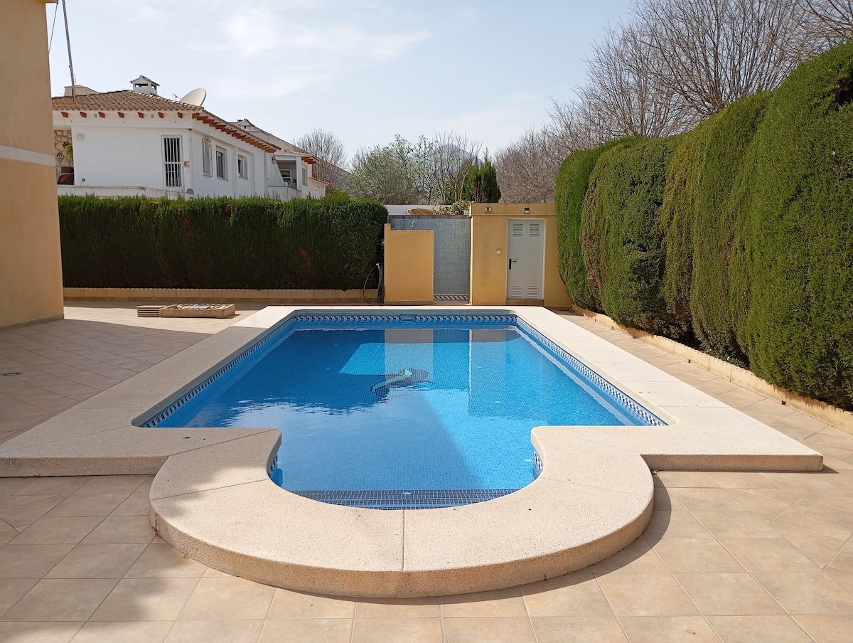A clear blue pool is surrounded by neatly trimmed hedges and a tiled patio. Sunlight reflects off the water's surface, highlighting the pool's inviting shape. A small privacy wall and a door are visible in the background, enhancing the sense of seclusion.