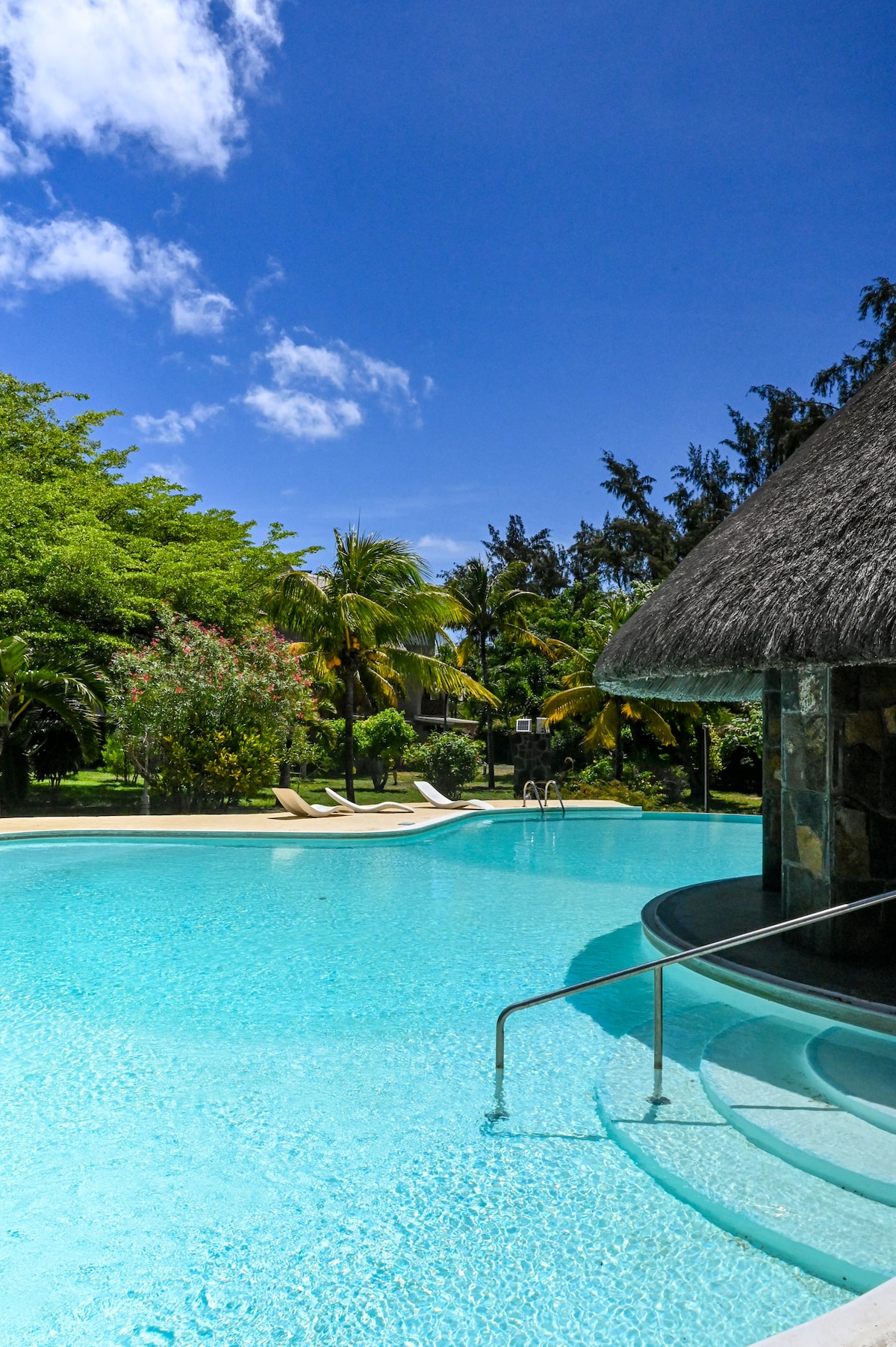 A large pool is set within a tropical garden, featuring clear turquoise water. Sun loungers are positioned next to the pool, surrounded by lush greenery and palm trees. A thatched-roof structure is visible nearby, providing shade and aesthetic charm.