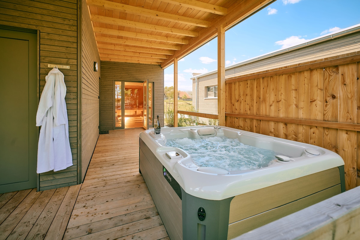 A serene outdoor area features a hot tub surrounded by wooden decking. Soft light filters through the open spaces, highlighting the natural wood elements. A bathrobe is hung nearby on a hook, adding to the relaxation ambiance.