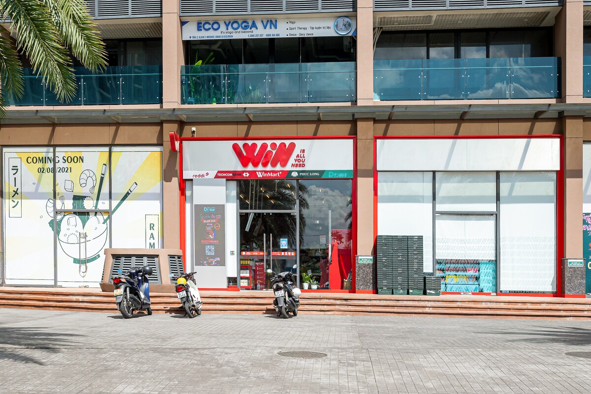 The image depicts a storefront labeled 'WIN' with large glass windows and red accents. Parked motorcycles are visible in front of the entrance, and a nearby advertisement indicates an upcoming opening. Surrounding palm trees add a touch of greenery to the scene.