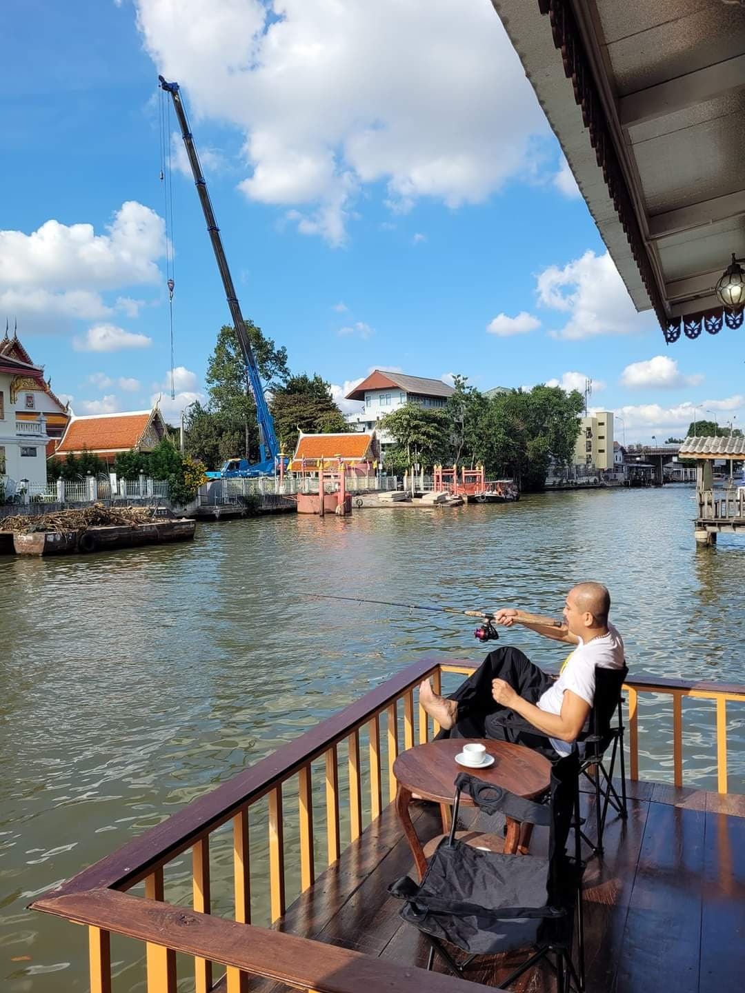 A person is seated on a wooden deck by the water, enjoying a fishing moment. A small table holds a cup, while a crane works in the distance against a backdrop of blue skies and scattered clouds. Nearby buildings and traditional architecture are visible across the river.