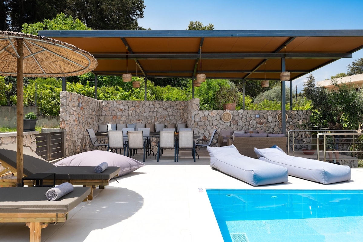 A well-designed outdoor area features a private swimming pool next to sun loungers and a shaded dining space. A large table surrounded by multiple chairs is placed under a covered structure, while natural stone walls provide a rustic backdrop to the serene setting.