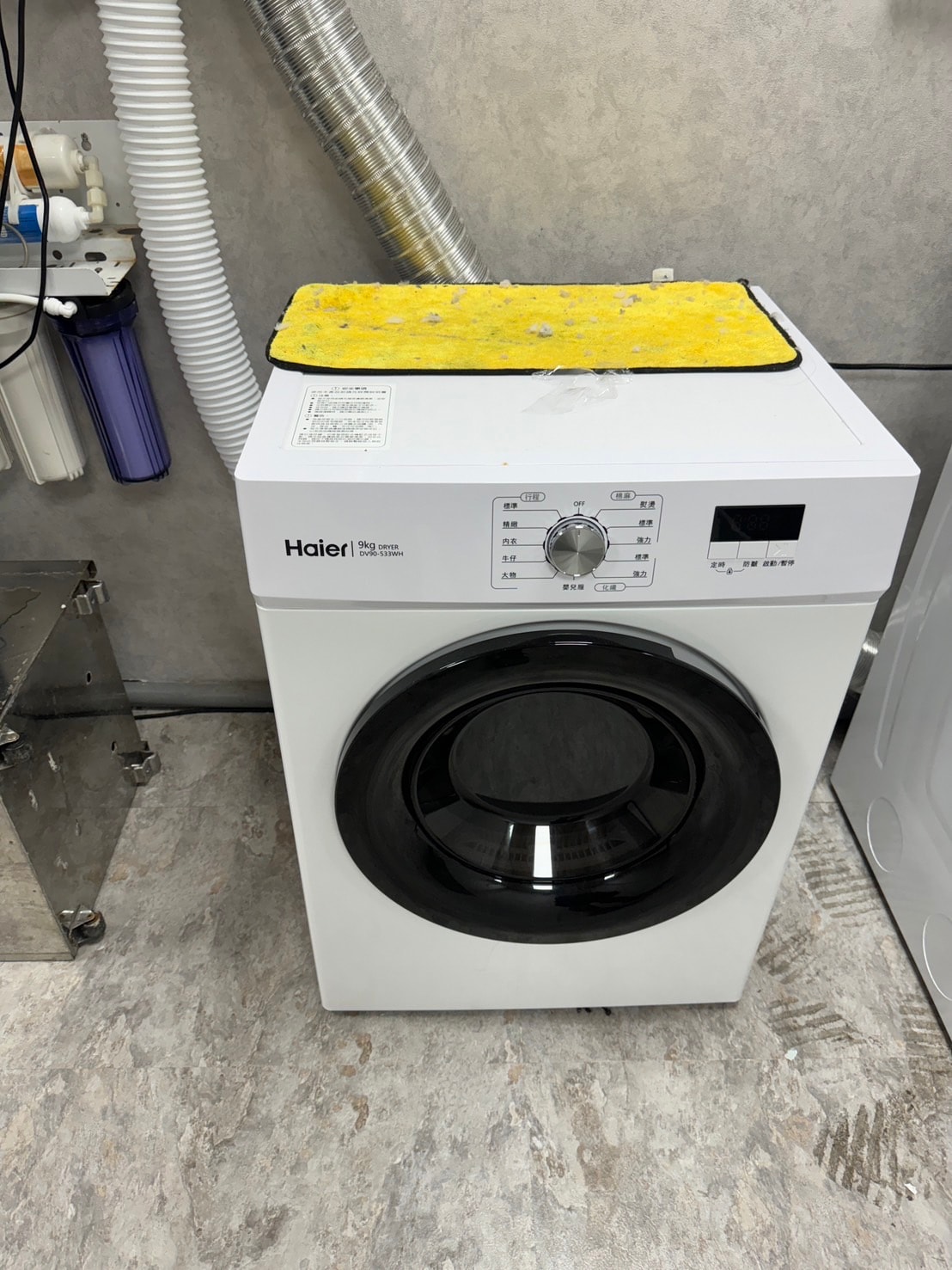 A front-loading washing machine is positioned against a textured wall. The machine features a round transparent door and a control panel with various settings. A yellow mat sits on top, providing a contrasting color to the overall white and gray tones of the laundry area.