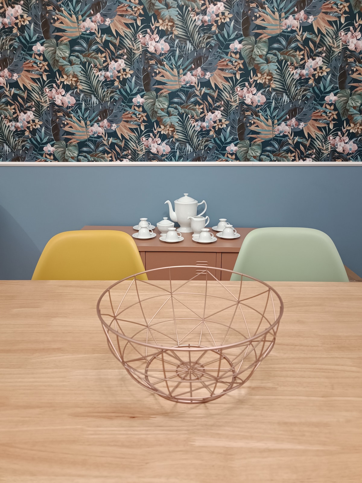 A modern dining area features a wooden table topped with a geometric wire fruit bowl. In the background, a colorful tropical mural contrasts with soft blue walls. A sideboard displays a complete tea set, ready for use, in harmonious white porcelain.
