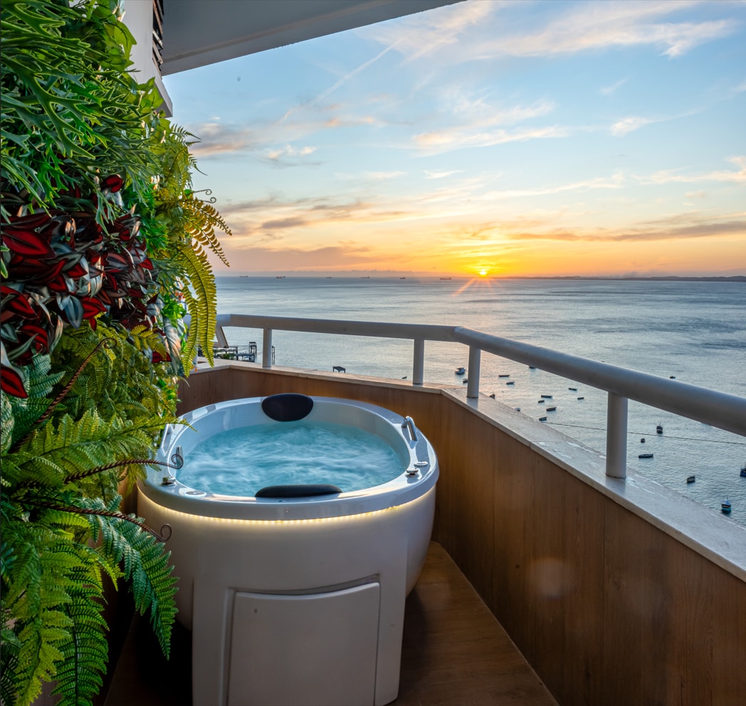 A private hot tub is positioned on a balcony, overlooking a calm bay at sunset. Soft jets of water create an inviting atmosphere, while greenery adds natural accents to the space. The horizon features a soft blend of warm colors as the sun sets beyond the water.