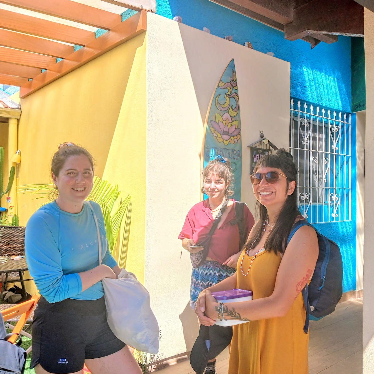 A vibrant outdoor area is depicted, featuring colorful walls in shades of yellow and blue. Several people are standing together, with surfboards displayed on the wall in the background. Comfortable seating is available, bordered by greenery that adds a relaxed vibe to the setting.