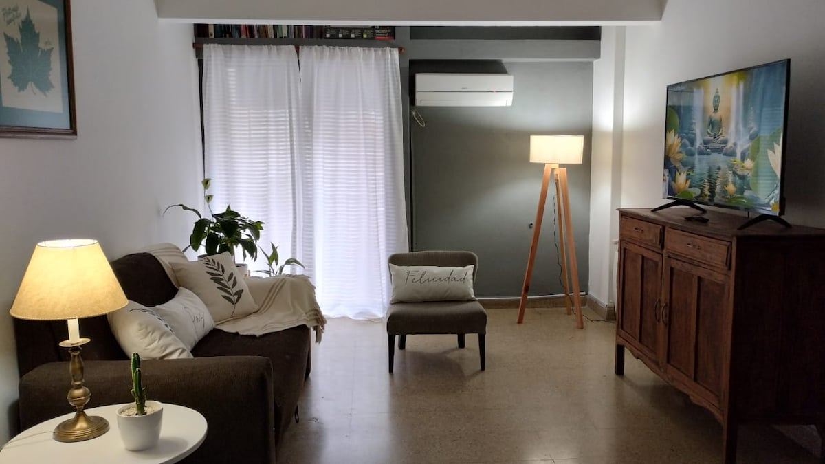 The living area features a comfortable sofa with light-colored accents, a stylish armchair, and a wooden cabinet. Natural light filters through sheer curtains, while a floor lamp and television contribute to the space's functionality.