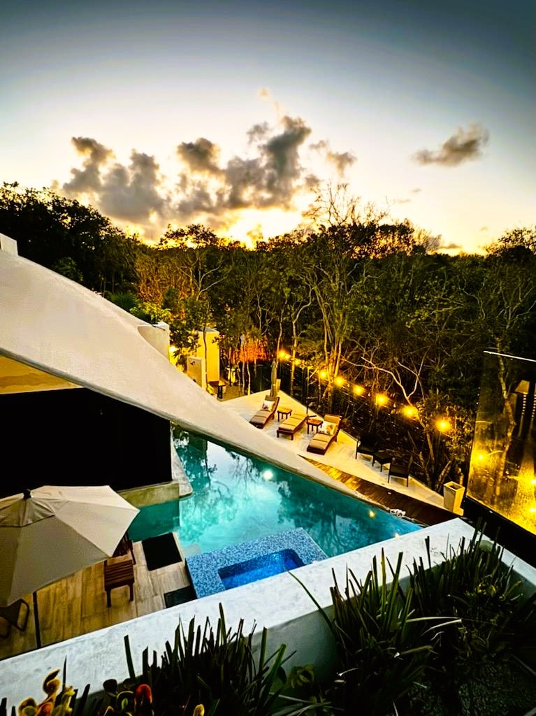 An aerial view captures a serene pool surrounded by lush greenery and illuminated by warm outdoor lights. Comfortable lounge chairs line the pool area, with an umbrella providing shade. The setting sun casts a gentle glow, enhancing the peaceful ambiance.